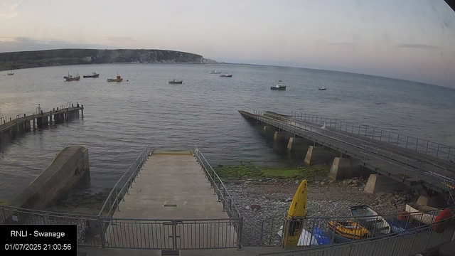 A view of a harbor at dusk, with several small boats floating on calm water, a dock extending into the water on the left, a sloped concrete ramp leading down to the shore, and a yellow caution sign near the bottom center. The background features a distant hilly coastline under a cloudy sky.