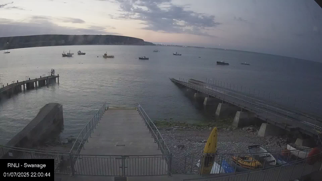 A view of a lighthouse pier extending into the water with two people standing at the end. Several boats are anchored across the water, with a distant shoreline and cliffs on the horizon. The sky is cloudy with the sun setting or rising, creating a soft, muted light. A concrete slipway and some parked vehicles are visible in the foreground.