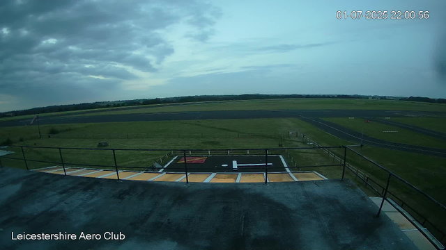 An expansive view of an airfield at Leicester Aero Club, taken from a webcam. The image shows a helipad in the foreground with a painted white "H" and a red square, surrounded by a railing. Beyond the helipad, there are grassy fields with a few small structures and a paved runway that extends into the distance. The sky is partly cloudy, with dark clouds on the left and patches of blue sky on the right. The timestamp in the upper right corner reads "01-07-2025 22:00:56."