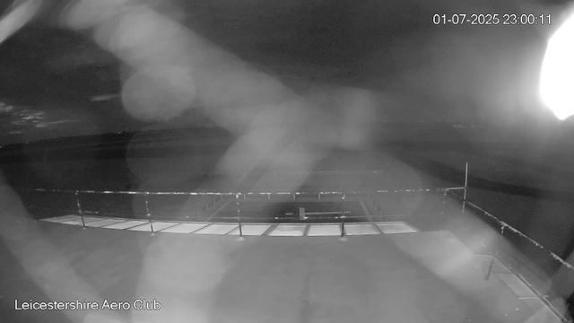 A black-and-white webcam image shows an empty outdoor helipad with a railing around it, captured at night. The sky appears dark, with a bright light source on the right side creating glare and lens flare effects. There is a timestamp in the top right corner reading "01-07-2025 23:00:11," and the bottom left corner displays the text "Leicestershire Aero Club."