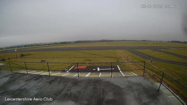 An overcast view from a webcam at Leicestershire Aero Club shows the airfield with a small helicopter landing pad in the foreground. The surrounding grassy areas and a taxiway or runway curve into the distance under a gray, cloudy sky. Faint hills can be seen on the horizon. The timestamp indicates the date and time as February 7, 2025, at 5:00 a.m.