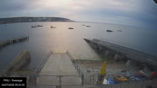 A calm seaside scene at dawn with a view of a small harbor. Several boats are anchored in the water, floating smoothly. There are two concrete piers extending into the water, one on the left and one on the right, with metal railings. The sky is mostly cloudy with a faint glow of sunlight breaking through, illuminating the scene softly. The timestamp indicates it's early morning on July 2, 2025.