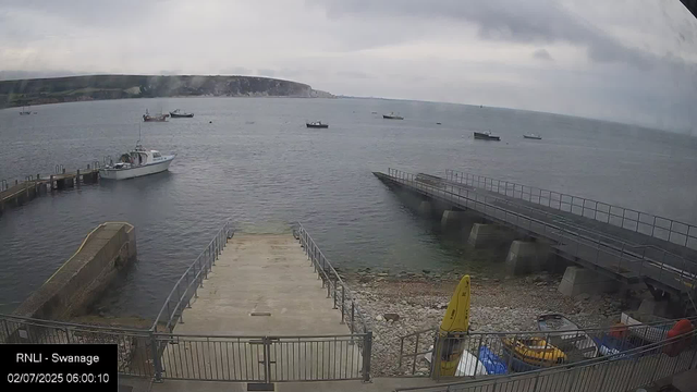 A view of a small harbor with several boats anchored in the water. There is a concrete dock extending into the sea, with some railings along its edges. On the right side, there is a sloped ramp with railings leading down to the water, and some equipment and yellow safety gear are visible near the bottom of the ramp. The sky is overcast with gray clouds, and distant cliffs line the horizon. The image has a timestamp of 02/07/2025 06:00:10 and is labeled "RNLI - Swanage."