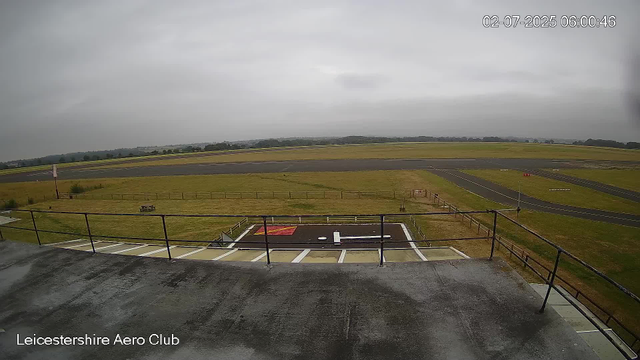 A wide view of an overcast sky above a runway and grassy airfield at Leicester Aero Club. The image is taken from an elevated position, showing the tarmac, a small helipad with a white helicopter landing mark, and the surrounding open fields. The date and time are displayed in the upper right corner.