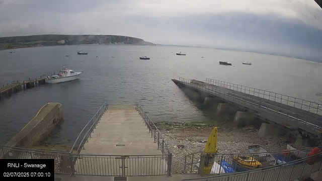 A view of a harbor with several boats anchored in the water. There is a concrete dock area in the foreground with a yellow buoy and some equipment on the rocky shore. The sky is cloudy, and a distant landmass with cliffs is visible on the horizon.