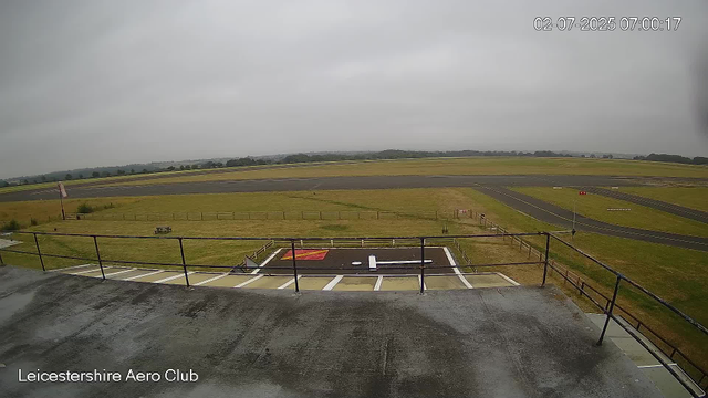 A view from a webcam at Leicestershire Aero Club shows an overcast sky with gray clouds. In the foreground, there is a weathered concrete platform with metal railings, and a helipad marked with a white "H" and a red border. Beyond the platform, there is a grassy area, a bench, and a small wind sock. The tarmac runway extends into the distance, with a few signs and markers along its edges. The landscape is flat, with a few trees and low hills visible on the horizon. The timestamp on the image indicates it was taken on July 2, 2025, at 7:00 AM.