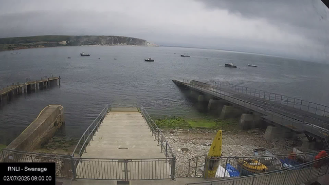 A view of a cloudy seaside scene with several small boats anchored on calm water. There are two concrete piers extending into the water, with a small boat on the shore near the bottom right corner. Green algae cover part of the shore, and a yellow buoy is standing upright. In the background, there are cliffs with a mix of white and dark rock, under a dark, overcast sky.