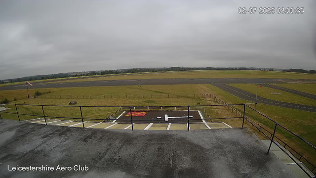 A view from a webcam at Leicesterhire Aero Club shows an overcast sky with gray clouds. In the foreground, there is a small helipad marked with a large "H" and a red and white landing area, surrounded by a safety fence. Below, the platform's surface appears wet. Beyond the helipad, there are grassy fields, a paved runway, and a few small structures. The scene is calm with no visible activity or vehicles. The image is timestamped "02-07-2025 08:00:35" in the top right corner.