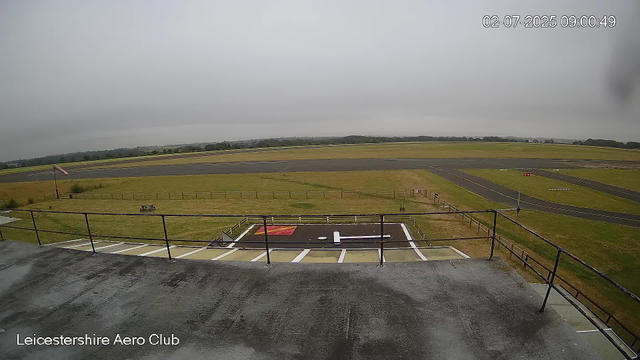 An overcast scene at Leicestershire Aero Club showing a helicopter landing pad in the foreground with white lines and a red marking. A black fence surrounds the pad, and there is a small tent-like structure on the grass nearby. Beyond the grass, runways extend into the distance under a cloudy sky. The image is timestamped February 7, 2025, at 9:00 AM.