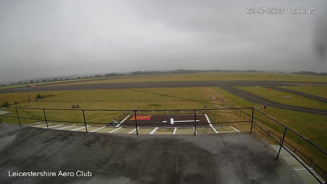 The image shows an overcast sky at Leicestershire Aero Club, with a weathered tarmac area in the foreground featuring a small helipad marked with a white 'H' and a red landing zone. Beyond, there is green grass enclosed by a wooden fence, and a small windsock on a pole indicating a light wind. In the background, there is a single runway with markings and a distant landscape of trees and hills, all under cloudy skies. The timestamp in the top right corner reads "02-07-2025 10:00:45."