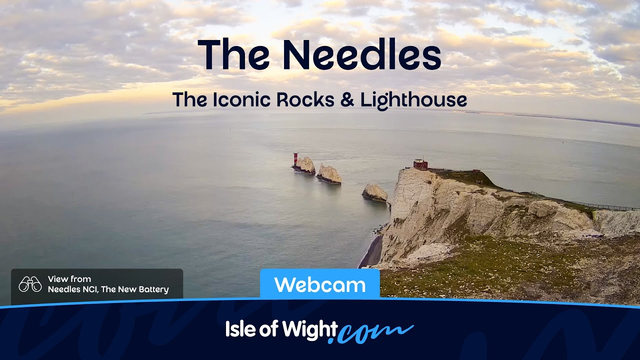 An aerial view from a webcam shows the iconic white chalk cliffs called the Needles on the Isle of Wight, standing in the sea with a lighthouse on a small rock formation nearby. The sky is partly cloudy with soft, golden light, suggesting either sunrise or sunset. The caption overlay reads "The Needles" and "The Iconic Rocks & Lighthouse," with additional text indicating the view is from Needle's NCI, The New Battery. The bottom of the image features the logo and website for Isle of Wight.com.