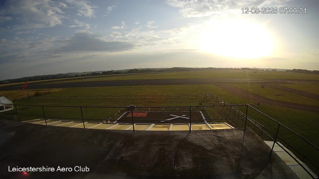 An outdoor view of the Leicester Aero Club featuring a grassy airfield with runways, a helipad with a red marking, and a surrounding fence. The sky is partly cloudy with the sun low on the horizon, indicating either early morning or late afternoon. There is a small tent and some equipment near the fence, and a few distant trees and fields extend into the background. The timestamp shows "12-08-2025 07:00:21".
