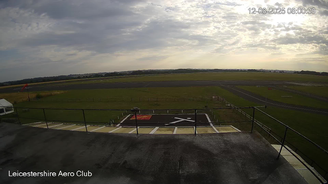 An empty helicopter landing pad with black and white markings, surrounded by a safety railing, is visible in the foreground. Beyond it, there is a grassy area with a windsock on the left side. In the background, an open airfield with runways and taxiways stretches into the distance. The sky is partly cloudy with sunlight breaking through the clouds. The timestamp in the upper right corner shows "12-08-2025 08:00:39" and the text "Leicestershire Aero Club" is visible at the bottom left corner.