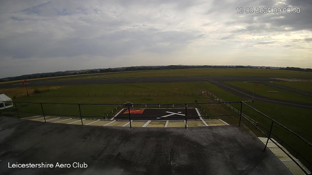 An image from a webcam showing a cloudy sky over an airfield at Leicester Aero Club. The view includes a helipad in the foreground, surrounded by a safety railing, and a runway stretching into the distance. There are some structures and grassy areas near the helipad, and the scene appears to be in the early morning. The date and time stamp reads "12-08-2025 09:00:30."