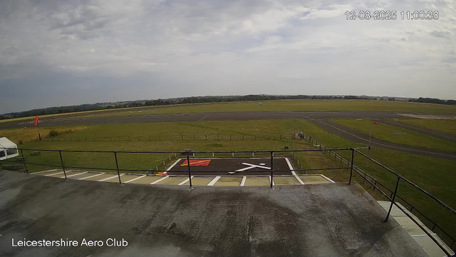 An empty helicopter landing pad is seen in the foreground, with a black and white X marking. Beyond the pad, there is a grassy area with a small fence, and several taxiways and runways extending across the airfield. The sky is partly cloudy, and the landscape stretches into the distance with trees and low hills. The image is timestamped "12-08-2025 11:00:38" in the top right corner, and "Leicestershire Aero Club" is written at the bottom left.