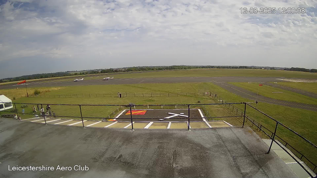 A view of an airport runway and taxiway from the perspective of a webcam at Leicesterhire Aero Club. In the foreground, there is a small helipad marked with a white "h" and a red and yellow pattern, enclosed by a railing. Below the camera's view, a group of people can be seen walking on a pathway near a tent. Two small airplanes are parked on the apron, with one flying in the background. The sky is mostly cloudy with patches of blue, and the timestamp in the top right corner reads "12-08-2025 12:00:08".