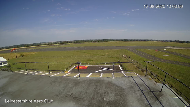 An empty tarmac area at Leicestershire Aero Club with a parking space and a helicopter landing pad marked with a white helicopter symbol. In the background, there's a runway with a windsock on the left side. The scene is outdoors under a partly cloudy blue sky, and the date and time are displayed in the top right corner as "12-08-2025 13:00:06."