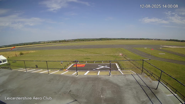 An outdoor view of an airport runway and taxiway area on a clear day with a blue sky and some scattered clouds. In the foreground, there is a rooftop observation deck with a railing, some benches, a marked helipad, and a person standing near the railing. A windsock is visible on the left side of the grassy area. The phrase "Leicestershire Aero Club" appears in the bottom left corner of the image. The date and time stamp on the top right corner shows "12-08-2025 14:00:08."