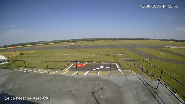 An outdoor view of a small airport or aero club facility under a clear blue sky. In the foreground, there is a fenced helipad with a red and white ground marker and a red windsock on a pole. To the left, there is a white tent and some benches. The runway extends into the distance with a few small aircraft visible. The landscape is flat with grassy fields, and the scene is labeled "Leicestershire Aero Club" at the bottom left corner. The timestamp shows "12-08-2025 14:59:55".
