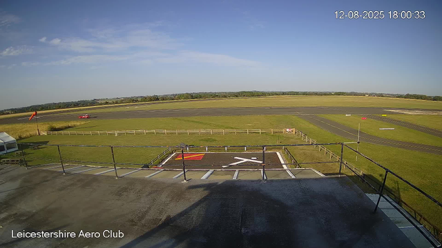 A view from a webcam at Leicester Aero Club showing a clear day with a blue sky and a few clouds. In the distance, there is a small red aircraft on the runway, with some runway markings visible. The foreground shows a balcony area with a railing and marked parking spaces below. The grassy airfield extends into the horizon with some trees in the far distance. The date and time displayed in the upper right corner is 12-08-2025 18:00:33.