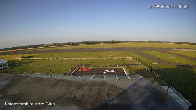 An outdoor view from a webcam at Leicestershire Aero Club shows a clear sky, a grassy area with a helipad marked with a white helicopter icon in the foreground, and a small white tent on the left side. The grassy field extends toward a paved area, likely part of a runway or taxiway, with various markings. In the distance, there are trees and open fields under a bright, sunny sky. The date and time are displayed in the upper right corner as "12-08-2025 19:00:29".