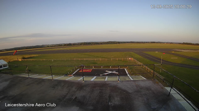 An empty helipad marked with a white helicopter symbol and a red square, surrounded by a metal railing, located on the rooftop of a building. Beyond the helipad, there is a vast open field with a short fence, and a paved runway extends into the distance under a clear sky during sunset or early evening. The image is labeled "Leicestershire Aero Club" in the bottom left corner, and the timestamp in the top right corner reads "12-08-2025 20:00:29".