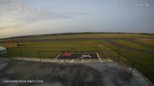 The image shows a view from a webcam at Leicestershire Aero Club, overlooking a small helicopter landing pad with a white helicopter symbol painted on the ground, surrounded by a safety barrier. Beyond the landing pad, there are green grassy fields and a runway in the distance under a partly cloudy sky. The timestamp in the top right corner indicates it was taken on December 8, 2025, at 21:00:34.
