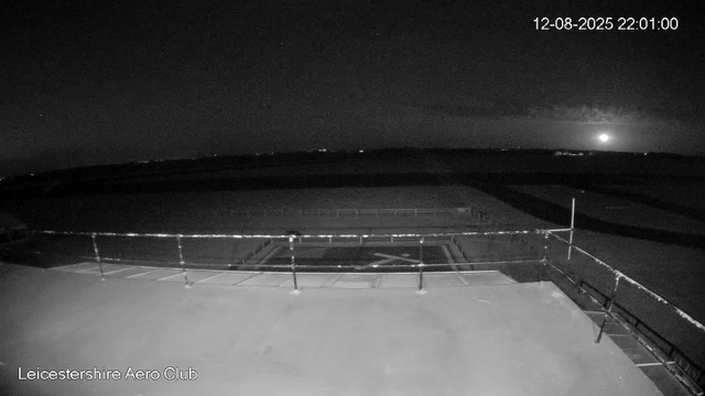 A black and white nighttime image showing an empty outdoor area at Leicestershire Aero Club. The scene includes a flat landing pad with a circular marking and a safety railing around its perimeter. In the background, a dark landscape stretches out under a starry sky, with a bright moon near the horizon on the right side. The timestamp at the top right corner reads "12-08-2025 22:01:00".