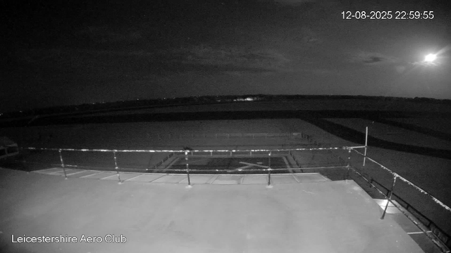 A black-and-white image from a webcam shows an empty rooftop area with safety railings around the edge. The sky is clear with a few clouds, and the moon is visible, casting light over the scene. In the distance, there are faint lights suggesting a distant landscape. The timestamp on the image is 12-08-2025 at 22:59:55, and the location is labeled as Leicesterhire Aero Club.