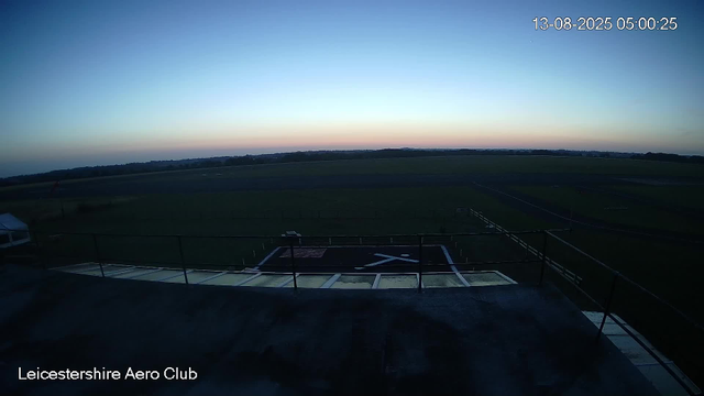 A view of the Leicesterhire Aero Club taken at dawn or early morning. The sky is clear with a faint gradient of colors from dark blue to light orange near the horizon. The foreground shows part of a rooftop terrace with safety railings, and a helicopter landing pad marked with a large 'X'. The surrounding landscape is flat, with open fields and a distant tree line on the horizon. The timestamp on the image reads 13-08-2025 05:00:25.
