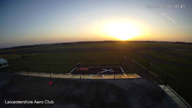 A sunrise over an airfield at Leicestershire Aero Club, with the sky gradually brightening and the horizon visible in the distance. The airstrip and surrounding grassy areas are visible in the foreground, with the club's helipad marked with a large white "H" on a red background. The timestamp indicates the date and time as August 13, 2025, at 6:00 am.