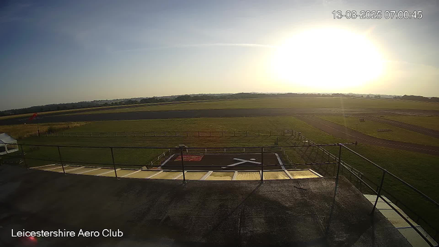 An early morning view from a webcam at Leicesterhire Aero Club. The sun is rising, casting a bright glow over the landscape. In the foreground, there is a small helipad with a white helicopter symbol, surrounded by a fence. Beyond that, there are open grassy areas and a runway extending into the distance. The sky is mostly clear with just a few wispy clouds. The timestamp shows the date and time as August 13, 2025, at 7:00 AM.