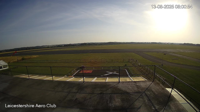 An outdoor view from a webcam at Leicesterhire Aero Club shows the early morning sky with the sun rising on the right side, casting bright light over the grassy airfield. There is a helicopter landing pad in the foreground surrounded by black railings, and a windsock on the left side near a small white tent. The runway and open fields extend into the distance under a partly cloudy sky. The timestamp in the upper right corner indicates it was taken on August 13, 2025, at 8:00 AM.