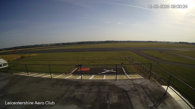 An outdoor view of Leicesterhire Aero Club showing a runway, grassy fields, and a helipad in the foreground. The sky is mostly clear with a few clouds and the sun shining brightly towards the top right. The image is timestamped 13-08-2025 at 09:00:24.