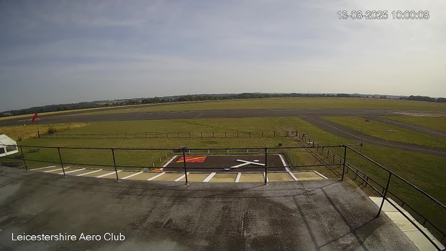 An empty tarmac area with a helipad marked with a white 'H' inside a black circle and outlined in white. Beyond the tarmac, there's a grassy field with a few small structures and a windsock on a pole. In the background, there are paved taxiways and runways, with a clear blue sky overhead. The image is timestamped "13-08-2025 10:00:08" and the location is noted as "Leicestershire Aero Club."