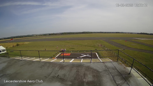 A view from a webcam overlooking a small aircraft runway and surrounding grassy fields. In the foreground, there is a helipad with white markings and a red rectangle, and a small fenced area with benches and people sitting on them. To the left, a white tent is partially visible. The sky is mostly clear with a few clouds, and the scene is well-lit. The timestamp and location "Leicestershire Aero Club" are shown in the top right corner.