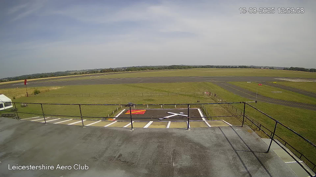 An empty apron area at Leicestershire Aero Club with a marked helipad, open grassy fields, a clear sky with some clouds, and the runway in the background. The timestamp shows the date and time as August 13, 2025, 12:59:58.