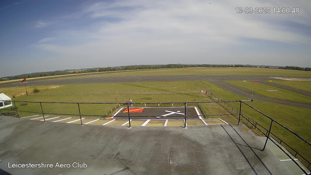 An image from a webcam at Leicestershire Aero Club shows a clear day with blue skies and some clouds. In the foreground, there is a small parking area with marked spaces, a fenced grassy area, and a person sitting on a bench. A windsock is visible near the grass, and the runway and open fields extend into the distance, with a tree line on the horizon. The timestamp indicates the photo was taken on August 13, 2025, at 14:00.