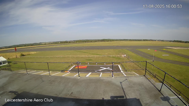 An outdoor view of an airfield at Leicestershire Aero Club under a clear blue sky. The foreground shows a helipad with a red and yellow mark, surrounded by a black metal fence, and a parking area with marked spaces. Beyond the fence, there is a grassy area and a short wooden fence, with a wide paved runway stretching into the distance. The landscape is flat and open, with fields and some trees on the horizon. The timestamp in the top right corner indicates the date and time as August 13, 2025, at 16:00:53.