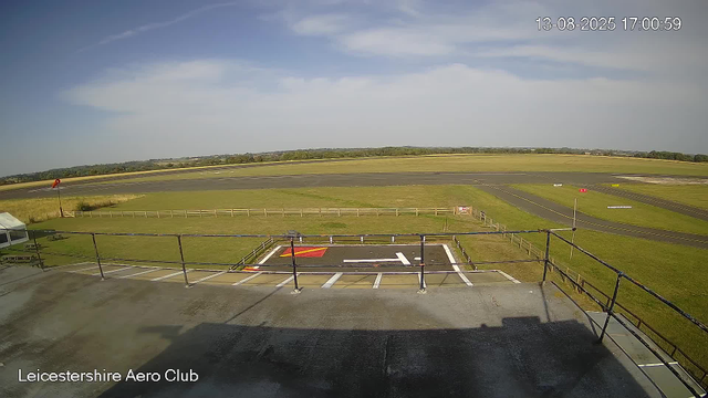 An empty view of an airfield taken from a webcam, showing a paved area with painted markings and a grassy field extending to a runway in the distance. The sky is partly cloudy with some patches of blue, and the scene is well-lit by sunlight. The timestamp in the upper right corner reads August 13, 2025, at 17:00:59. The bottom left corner displays the text "Leicestershire Aero Club."