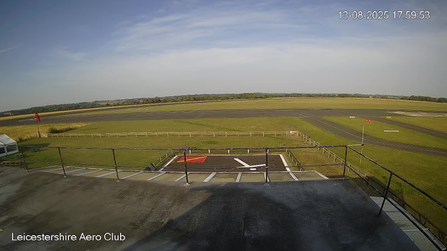 A wide view of an airfield taken from a webcam. The foreground shows a small helipad with a white helicopter symbol and red markings. There is a fenced area and some equipment on the left side. Beyond the fence, there are grassy fields and a runway extending into the distance. The sky is mostly clear with a few wispy clouds. The timestamp in the top right corner reads "13-08-2025 17:59:53," and the bottom left corner has the text "Leicestershire Aero Club."