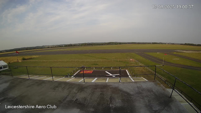 An image from a webcam showing an airfield runway with a small helipad in the foreground, marked with a red and white symbol and a white helicopter icon. There is a grassy area with a fence beyond the helipad, and the runway extends into the distance under a partly cloudy sky. A windsock is visible on the left side of the image, and a small white tent is partially visible on the left edge. The bottom left corner of the image has the text "Leicestershire Aero Club." The date and time displayed in the upper right corner are "13-08-2025 19:00:37."