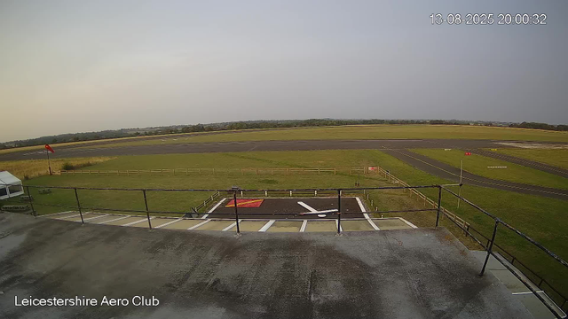 A view from a webcam at Leicestershire Aero Club showing an airfield with a paving area in the foreground, a grassy area, a windsock on a pole, and a clear sky. The runway extends into the distance with some markings, and a line of trees is visible on the horizon. The timestamp in the top right corner reads "13-08-2025 20:00:32."