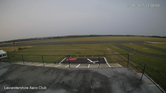 A view from a webcam at Leicester Aero Club shows an overcast sky, a grassy area with a taxiway and runway in the distance, and a helipad marked with a red and white pattern in the foreground. The scene is captured from a building balcony with a metal railing, and part of a small tent or canopy is visible to the left. The timestamp indicates the image was taken on August 13, 2025, at 21:00:01.