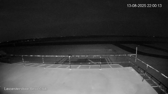 A black-and-white image captured by a webcam shows an empty outdoor area at night with a sports field surrounded by a metal fence. The scene is mostly dark, with some faint lights visible in the distant background. The date and time stamp in the upper right corner reads "13-08-2025 22:00:13," and the lower left corner has the text "Leicestershire Aero Club." The platform appears to be a rooftop or viewing deck with railings around the edge.