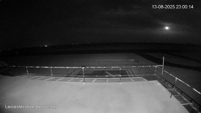 This black-and-white image from a webcam shows the Leicester Aero Club at night. The scene includes a parking area with empty spaces in the foreground, surrounded by safety railings. In the background, the sky features a bright moon and some scattered stars. The timestamp in the top right corner reads "13-08-2025 23:00:14."