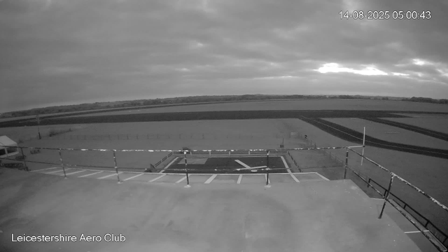 A black-and-white image from a webcam shows the tarmac and runway area of LeicesterShire Aero Club. The sky is cloudy with some sunlight breaking through. In the foreground, there is a parking area with painted lines and a small helipad marked with an "H." There are safety barriers along the edge of the parking area. In the distance, open fields and a horizon line are visible below the cloud-covered sky. The timestamp on the image indicates the date as August 14, 2025, and the time as 05:00:43.