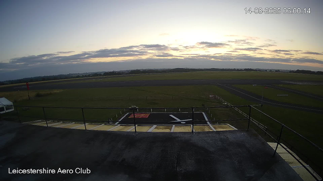 An early morning view of an airfield at the Leicestershire Aero Club, showing a runway, grassy areas, and a small platform with safety railings in the foreground. The sky is partly cloudy with the sun rising, casting a soft light over the scene.