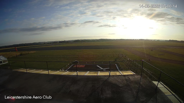 An outdoor view from a webcam at Leicesterhire Aero Club shows a grassy airfield with runways and taxiways stretching into the distance. The sky is partly cloudy with the sun low on the horizon, creating a bright glare. A small structure with a railing is in the foreground, and a person is standing near the edge of the runway area. The timestamp on the top right indicates the date and time as August 14, 2025, at 7:00 AM.