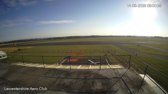 A wide view of an airfield at Leicester AerO Club on a sunny day. The foreground shows a small rooftop area with a red and white windsock on the left, a fence, and a helicopter landing pad marked with a red square and white outline. Beyond the fence, there's a grassy field, a paved runway, and a long stretch of open land with trees in the distance. The sky is mostly clear with a few scattered clouds, and the sun is shining brightly. The timestamp on the top right shows August 14, 2025, at 8:00 am.